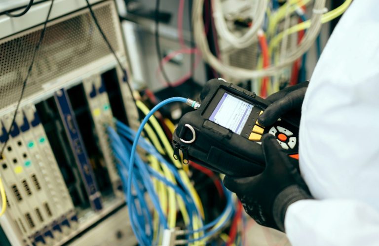 Eine Person hält ein Messgerät in der Hand und steht neben Server in einem Technikraum.